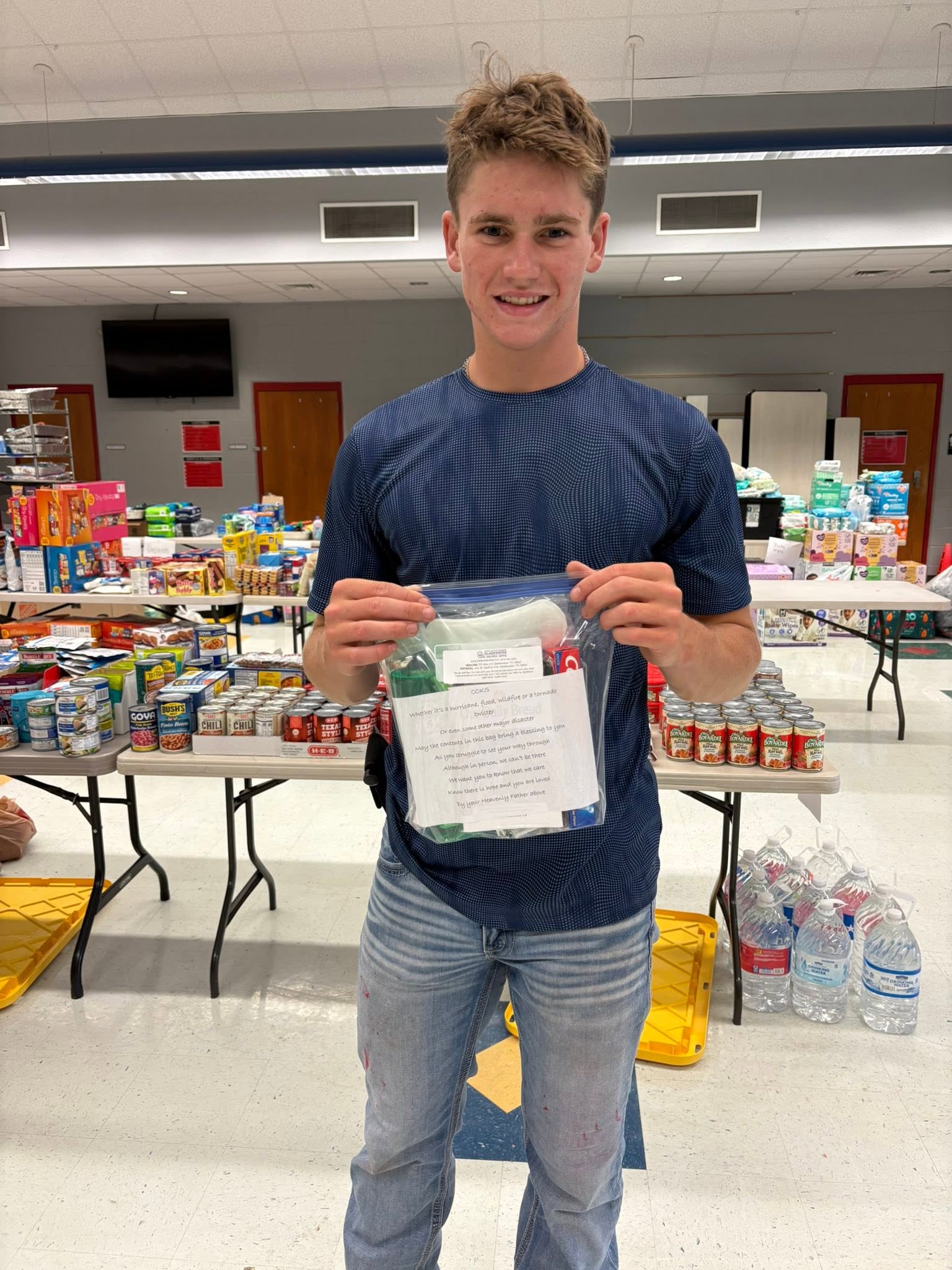 A community helper holds a relief kit beside tables of donated food and water in Kerrville.